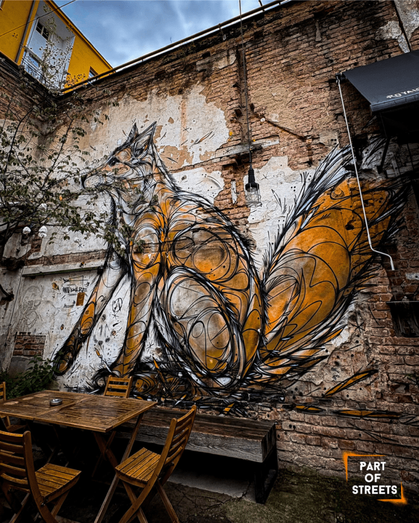 Mural of an abstract orange fox on a brick wall above wooden tables and chairs in an outdoor courtyard.