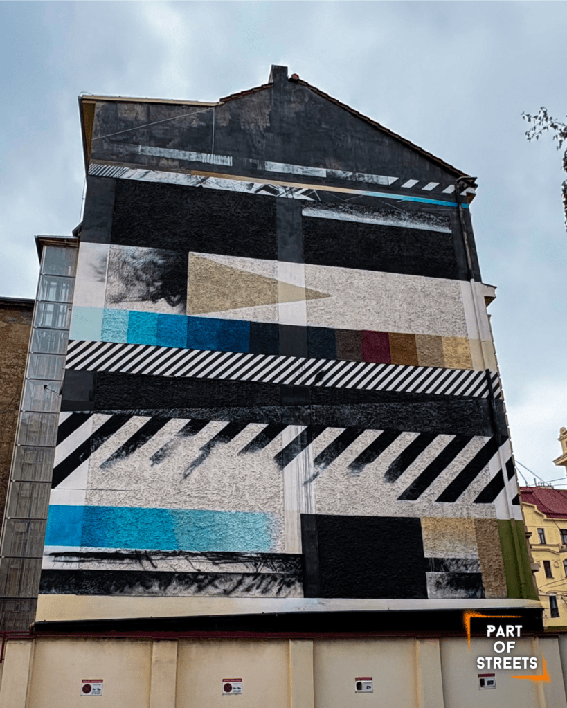 Abstract geometric mural with bold stripes, blocks of color, and patterns on the side of a tall building, with a white van parked below.
