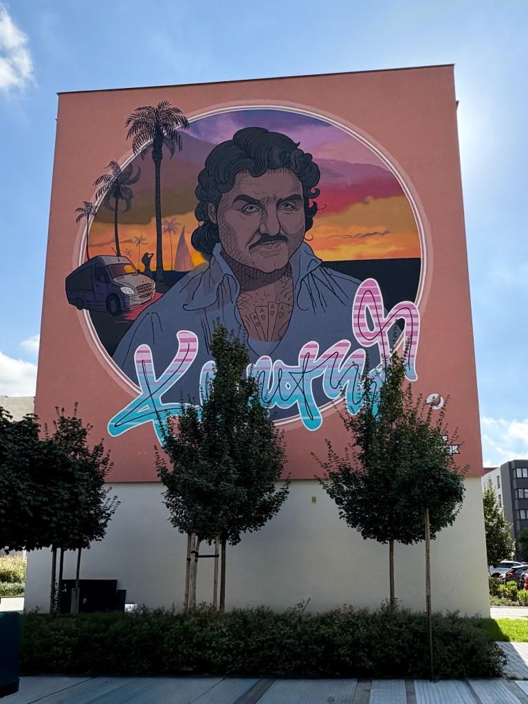 Mural of Krzysztof Krawczyk in Opole, painted by Bruno Neuhamer on Krakowska 34A. The artwork shows the Polish singer against a vivid sunset background with palm trees and a van, styled in retro Miami Vice colors with neon lettering of his name.