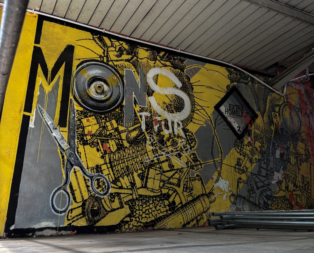 Industrial-style mural in Opole featuring bold yellow and gray tones, large scissors, abstract mechanical patterns, and the word “MONS” with stencil lettering.
