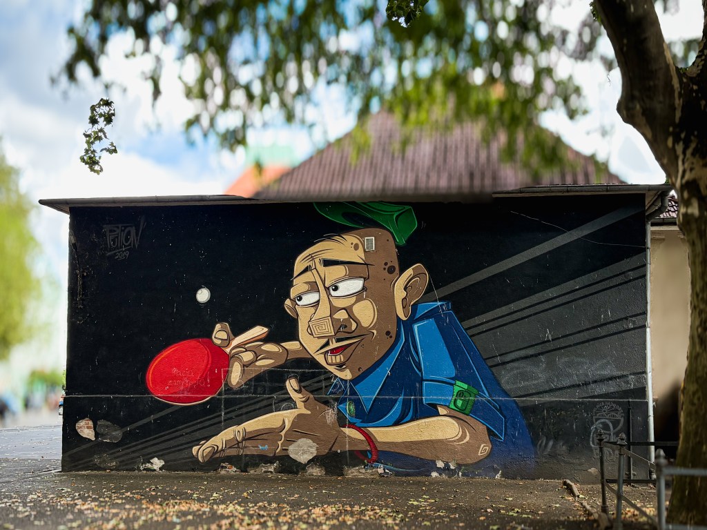 A mural on a dark wall depicting a cartoon-style man playing table tennis. He is wearing a blue shirt and a green cap, concentrating as he hits a small white ball with a bright red paddle. The background is black with diagonal gray lines.