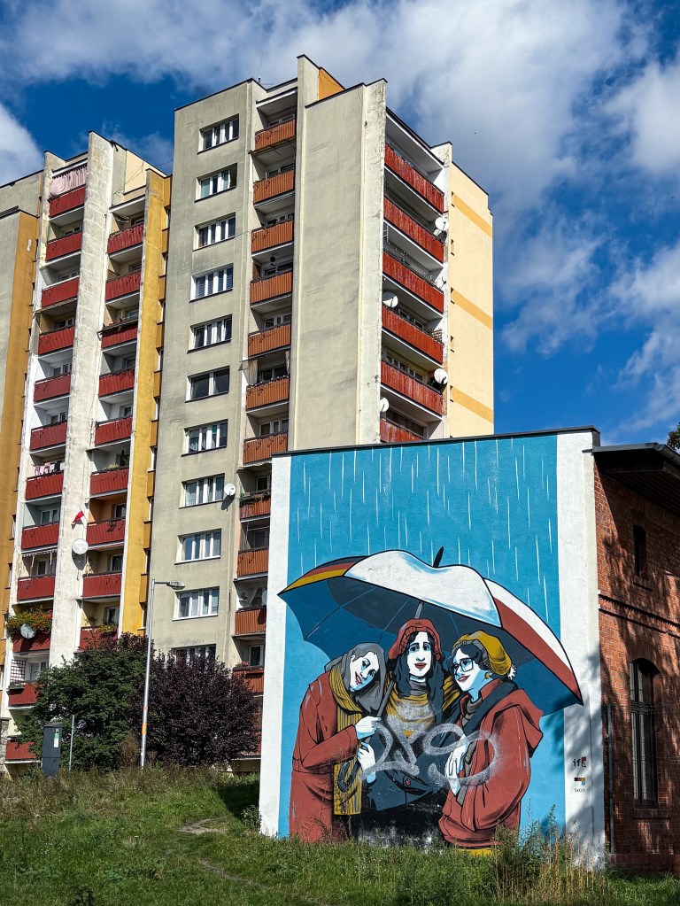 A colorful mural on the side of a small building depicting three women standing close together under a large umbrella in the rain. They are smiling and wearing scarves and coats. The background is blue with vertical white rain lines. The mural is located next to a tall residential apartment block with red balconies.