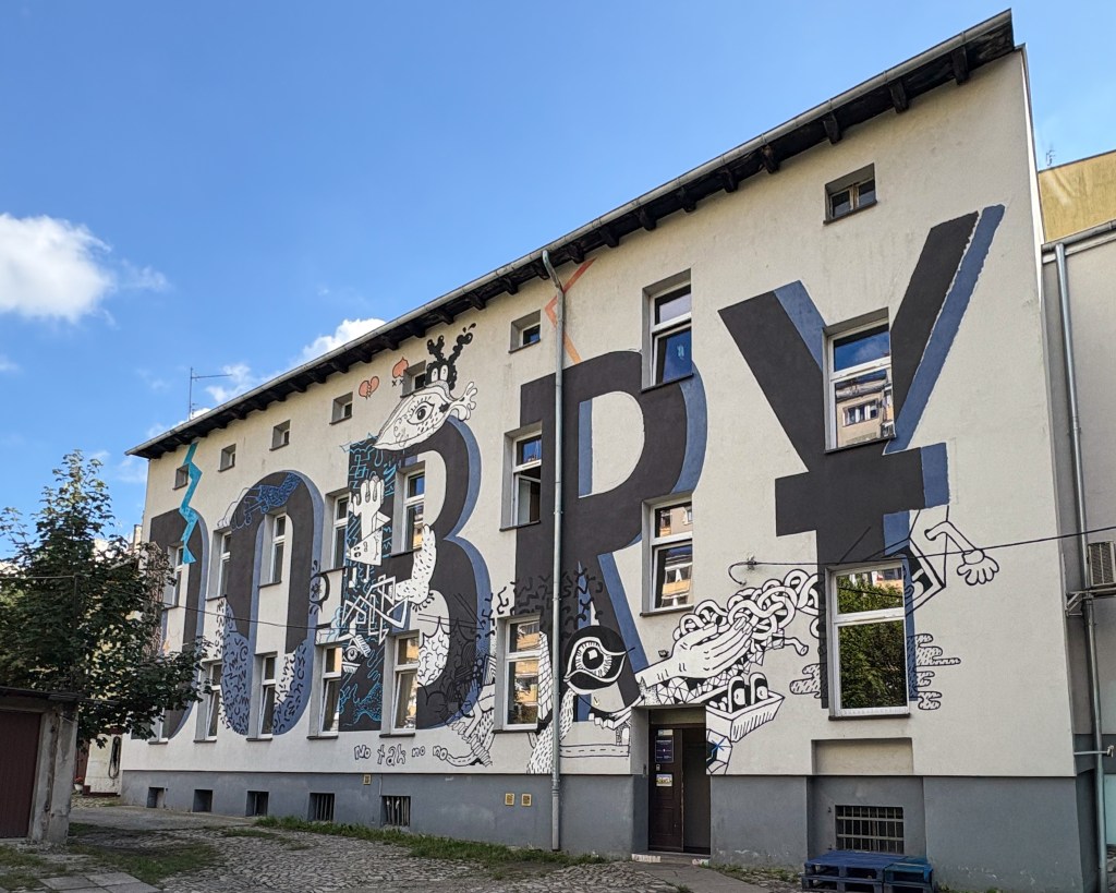 A colorful mural on a building façade in Opole featuring large, bold letters spelling “DOBRY” integrated with playful, surreal black-and-white illustrations of eyes, hands, and abstract shapes. The artwork combines geometric typography with imaginative doodles, set against a clear blue sky and cobblestone courtyard.