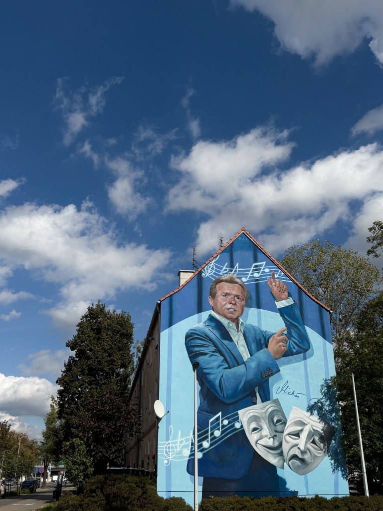 A large mural by Natalia Rak painted on the side of a tall building in Opole, Poland. It depicts a man in a blue suit conducting music, surrounded by floating musical notes and theatrical masks — one smiling, one sad — symbolizing art and performance. The mural stands out vividly against a bright blue sky with scattered clouds.