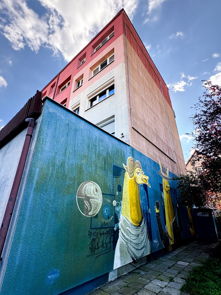 A colorful mural painted on the side of a low building next to a taller apartment block. The artwork features two stylized yellow llama-like characters dressed in clothing reminiscent of Star Wars, with one resembling Princess Leia and the other Han Solo. A small depiction of the Death Star floats nearby on a teal-blue background. The building behind the mural is painted in shades of pink, beige, and white.