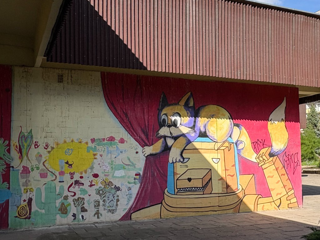 A colorful mural painted under a building overhang shows a large cartoon-style fox with big eyes and a bushy tail lying playfully on top of a robot-like box with a mouth full of teeth. The background features a red curtain pulled aside, revealing a section filled with childlike drawings — suns, animals, handprints, and abstract shapes — in bright colors like yellow, green, and pink. The wall and pavement below show signs of age, giving the scene a charming, urban feel.