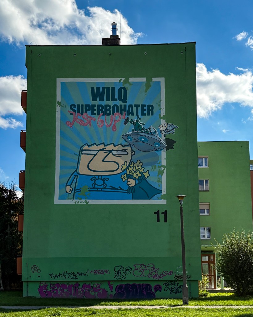 A large green apartment building with a mural of the Polish comic superhero WILQ. The mural shows a cartoon-style character with a serious expression, wearing a blue superhero outfit and holding flowers, with a flying saucer and other small comic figures in the background. The mural reads “WILQ SUPERBOHATER” (WILQ Superhero) and has some graffiti over it. The building number “11” is visible below the mural.
