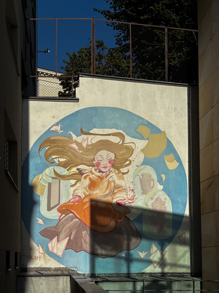 A colorful mural by Lisek.art in Opole’s city center, depicting a dreamy female figure with flowing hair and closed eyes, floating in front of a blue circular background. She’s wearing an orange and brown outfit, surrounded by soft shapes like folded paper cranes, letters, and bottles, giving the artwork a whimsical, serene atmosphere.