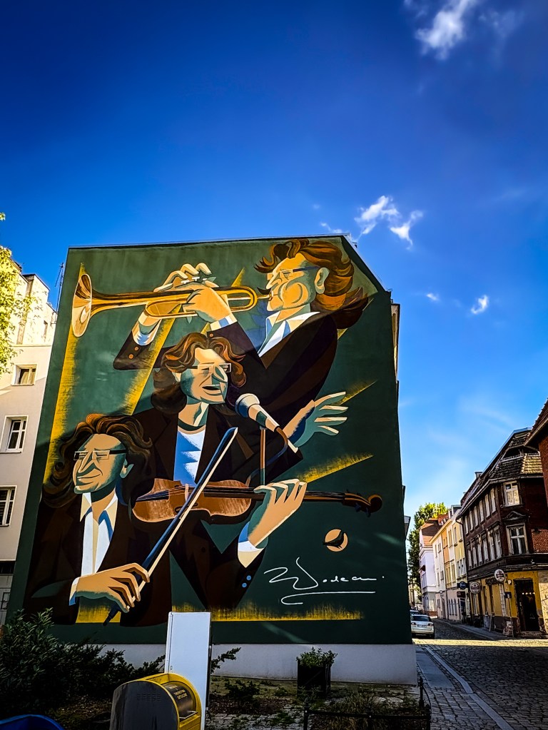 A vibrant mural in Opole depicting Zbigniew Wodecki - a musician performing in three dynamic poses — playing the trumpet, singing into a microphone, and playing the violin. The figure, with flowing brown hair and dressed in a suit, is painted in warm tones against a deep green background. The signature of the artist is visible at the bottom right corner of the mural.