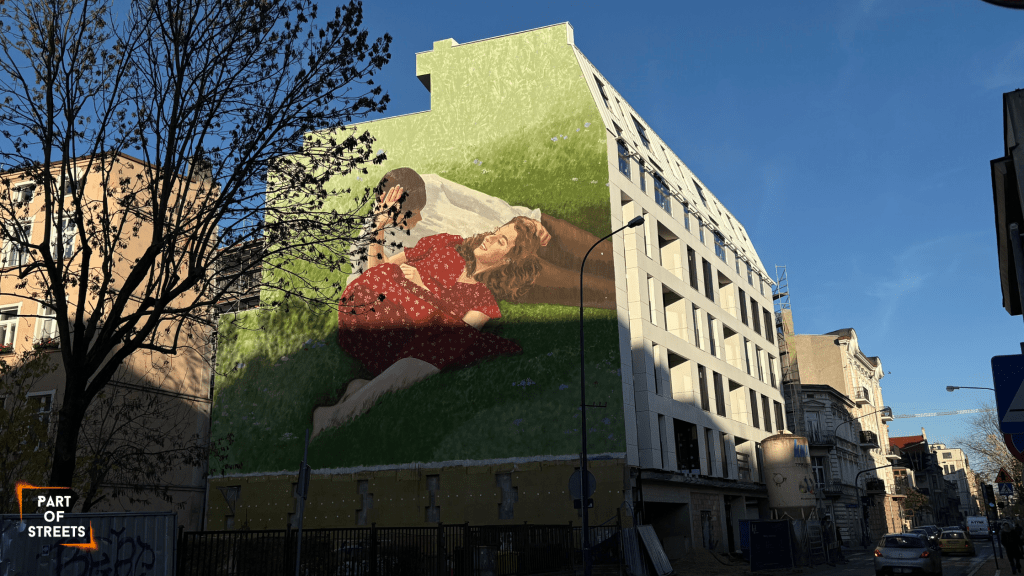 Street art mural in Łódź by Polish artist Paulina Kwietniewska, showing a woman in a red dress lying on green grass and resting on a white pillow. Large-scale mural painted on a building wall, part of the vibrant Łódź street art scene.
