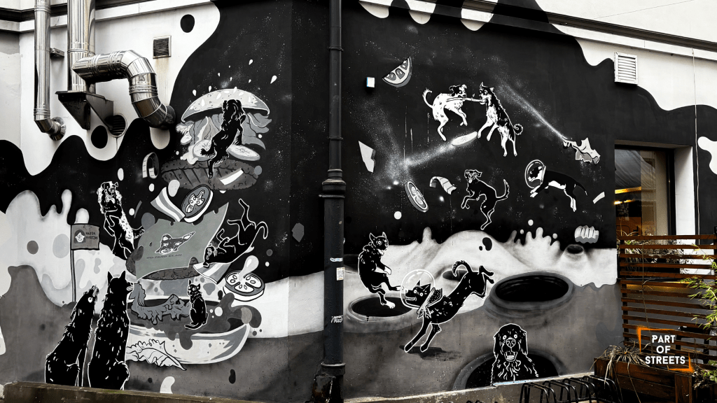 Black-and-white mural in Łódź featuring dynamic, cartoon-style dogs floating through a surreal space with food elements and abstract shapes. 