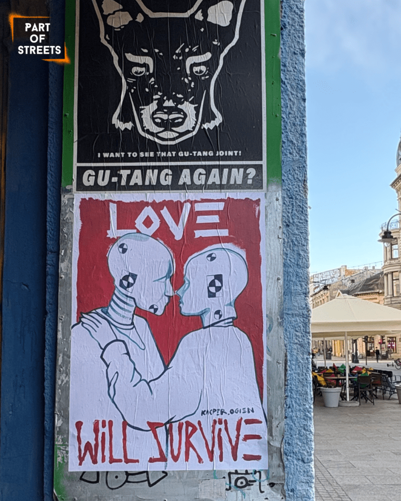 Street art poster in Łódź showing two simplified, humanoid robotic figures embracing against a red background. Their heads feature crash-test–style markings, and above them the word “LOVE” is written in bold red letters. At the bottom, handwritten text reads “WILL SURVIVE” with the signature “Kacper Ogień.” The poster is pasted on a column next to another black-and-white artwork featuring a dog and the text “GU-TANG AGAIN?”, with a city street and outdoor café visible in the background.