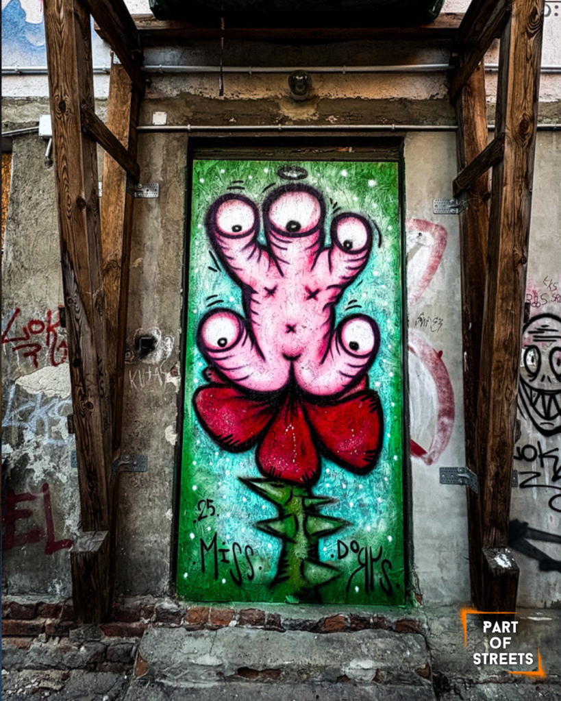 Colorful street art mural in Łódź depicting a surreal, five-eyed pink creature shaped like a flower, with red petals and a green thorny stem. The artwork is painted on an old doorway surrounded by weathered walls, wooden supports, and graffiti tags. Signed “Miss Dorys,” it blends vibrant cartoon style with the rough texture of an urban courtyard.