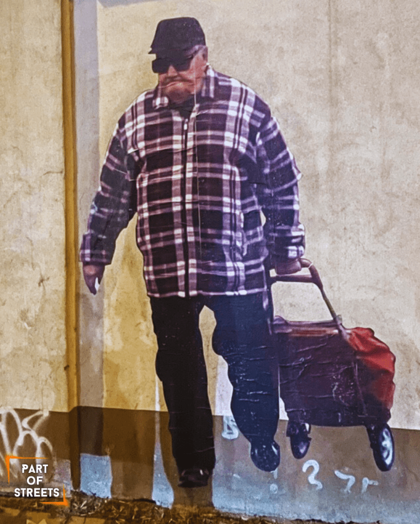 Street art paste-up in Łódź showing an older man walking with a red rolling shopping bag. He wears a black cap, dark sunglasses, and a large plaid jacket. The realistic figure is placed on a beige wall with faint graffiti at the bottom. Urban night lighting highlights the texture of the artwork and the rough surface of the building.