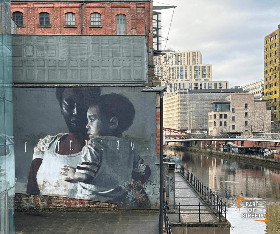 “Peterloo” mural by Axel Void in Manchester depicting a mother holding a child, painted on a riverside industrial building along the canal.
