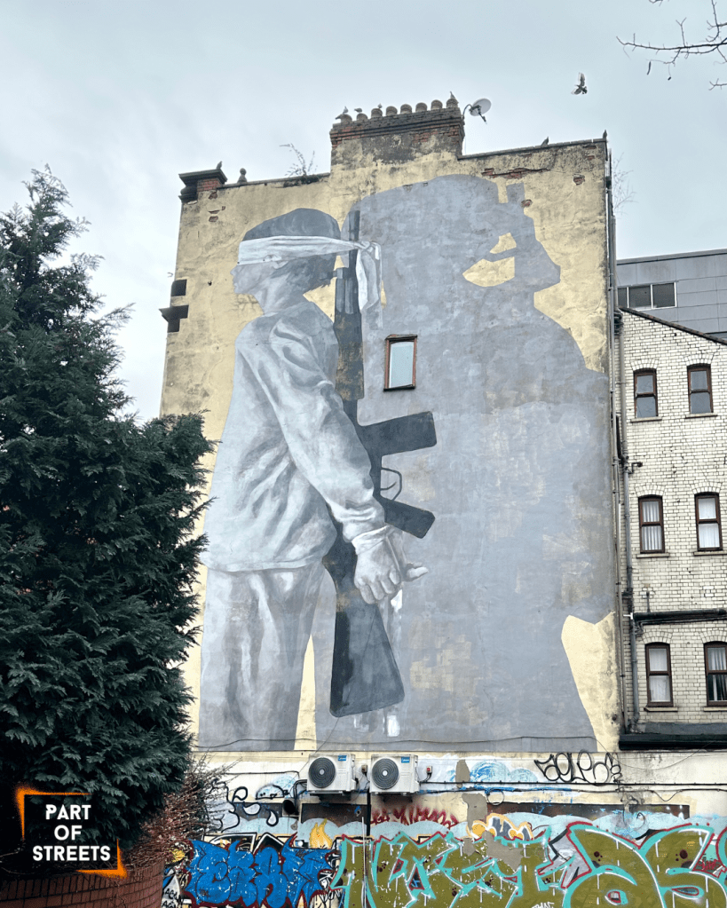 Large mural by Hyuro in Manchester showing a blindfolded child holding a rifle, painted in soft grey tones on a worn building facade, with the child’s shadow forming the silhouette of an armed adult figure — a powerful street art piece addressing lost innocence and conflict.