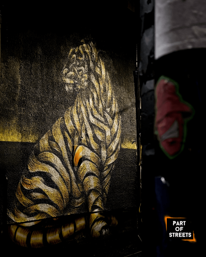 Mural by Otto Schade in Manchester depicting a striped tiger painted in the artist’s signature ribbon style on a dark urban wall.