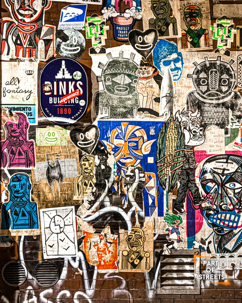 Street art paste-up wall in Manchester’s Northern Quarter covered with colourful posters, stickers, and urban artworks layered on a brick wall.