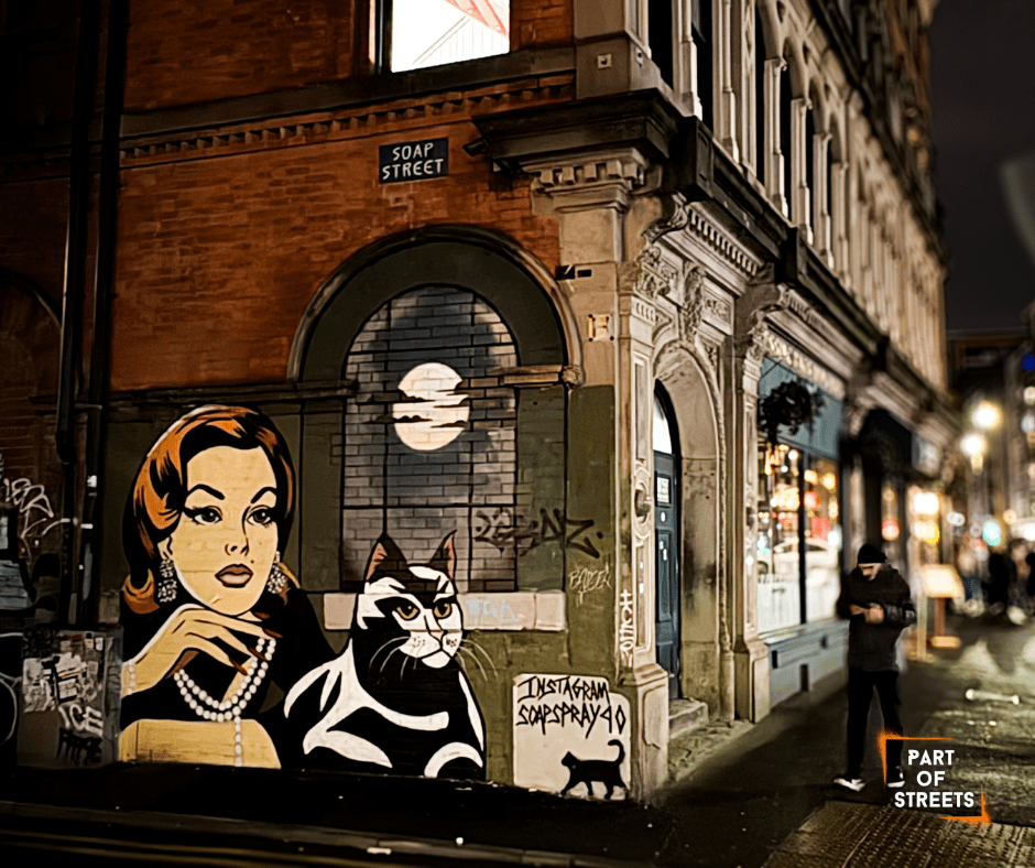 Street art by Soap Spray in Manchester featuring a retro-style female portrait and a black-and-white cat painted on a corner building on Soap Street at night.