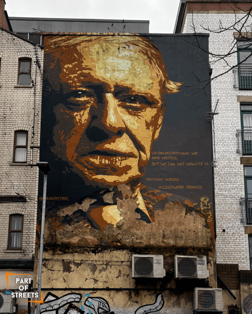 Large portrait mural by Tankpetrol in Manchester depicting writer Anthony Burgess in warm brown and golden tones, painted on a weathered building facade, accompanied by a quote from A Clockwork Orange.